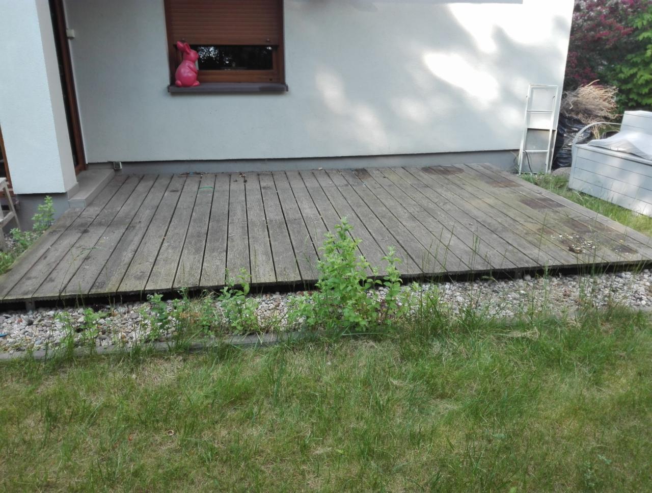 Drewniany taras przy białym budynku z widocznymi śladami zabrudzeń, otoczony kamieniami i zielenią, z różowym zajączkiem dekoracyjnym na parapecie okna.