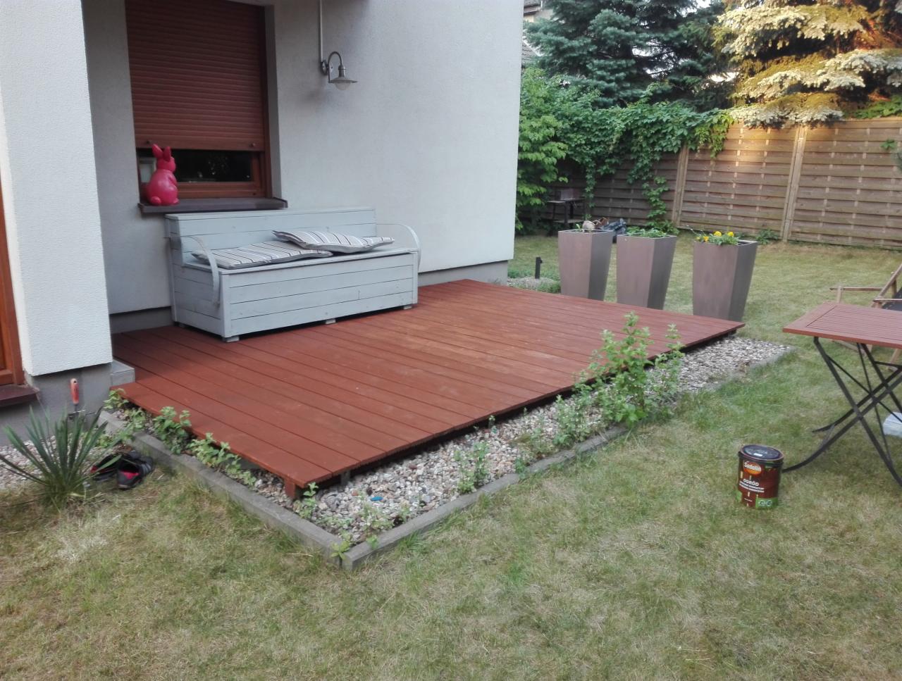 Świeżo pomalowany, brązowy taras drewniany przylegający do domu z szarą ławką i różowym króliczkiem na parapecie, obok widoczna puszka z farbą.