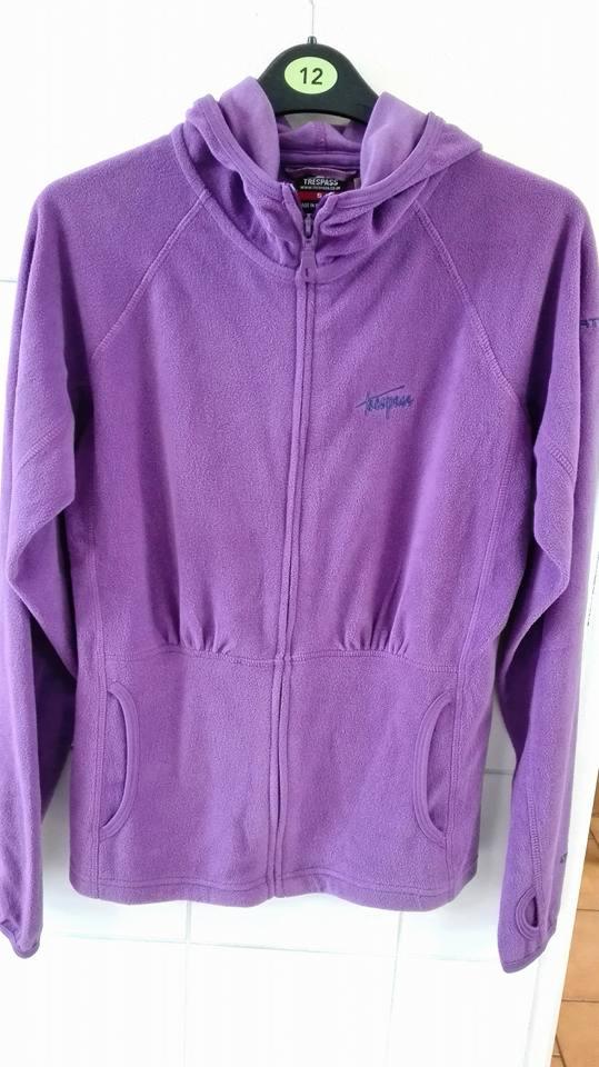 Fioletowa bluza polarowa marki Trespass z kapturem, zapinana na zamek, rozmiar 12 widoczny na wieszaku, z elastycznym ściągaczem w pasie i kieszeniami.