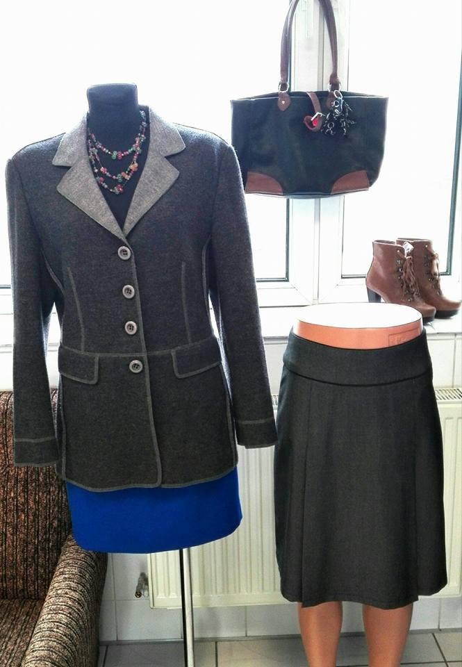 Szary damski żakiet z kontrastową lamówką i spódnica, prezentowane z torebką i botkami przy oknie.