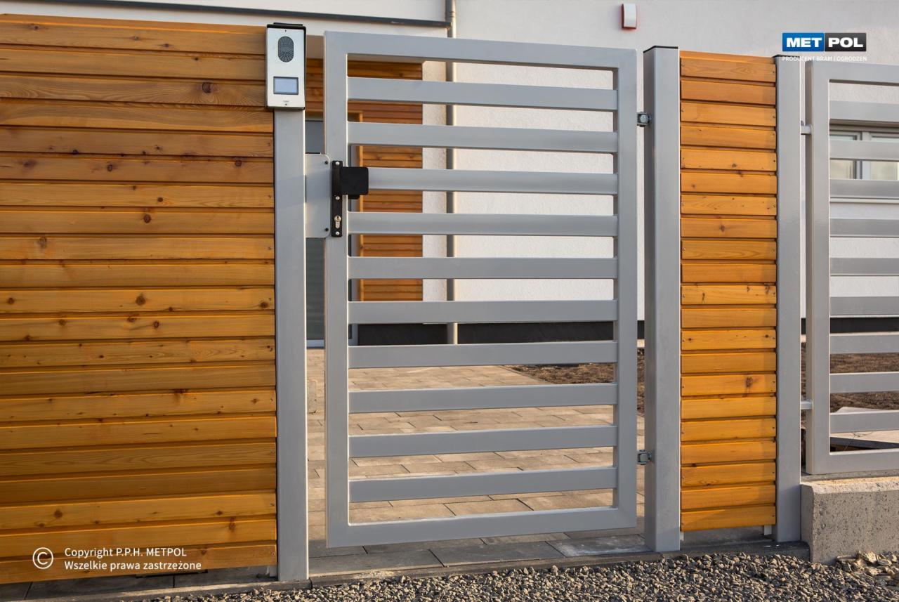 Nowoczesna, otwarta brama metalowa z poziomymi szczeblami w kolorze szarym, zamontowana na drewnianym ogrodzeniu z desek w odcieniach brązu, z widocznym panelem domofonu przy słupku.