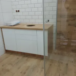 Biała szafka łazienkowa z drewnianym blatem pod umywalkę, widok na kabinę prysznicową w tle.