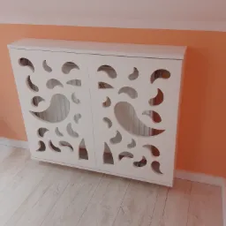 Biała, ażurowa obudowa grzejnika z motywem łezki, zamontowana na pomarańczowej ścianie, widok z lekkiego dołu.