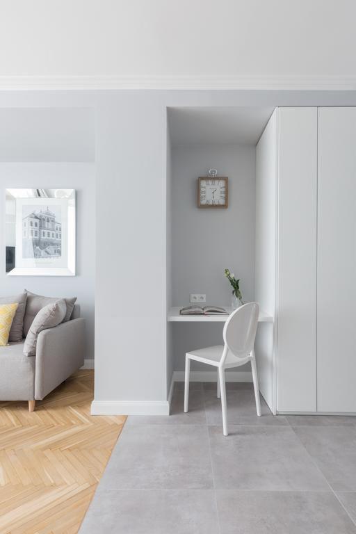Wnęka z biurkiem i białym krzesłem w minimalistycznym wnętrzu, obok szafa w zabudowie, na ścianie zegar, na biurku książka i wazon z kwiatami, w tle kanapa z żółtą poduszką i obraz w ramie.