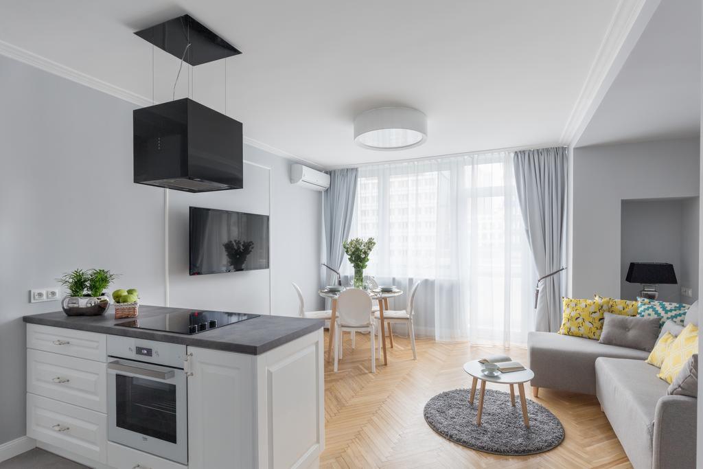 Jasne wnętrze mieszkania z aneksem kuchennym, białymi meblami, szarymi ścianami i parkietem w jodełkę; czarny okap nad płytą grzewczą, stół z kwiatami i szara sofa z kolorowymi poduszkami.