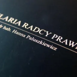 Tabliczka informacyjna z napisem 'Kancelaria Radcy Prawnego dr hab. Hanna Paluszkiewicz' na ciemnym tle, widoczna pod kątem.