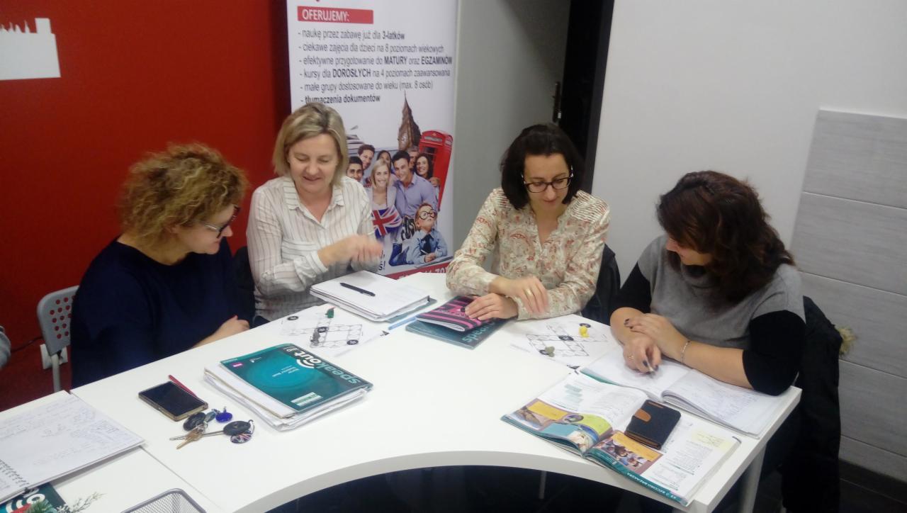 Cztery kobiety siedzą przy białym stole, pracując z podręcznikami i notatkami, w tle widoczny plakat z ofertą szkoły językowej.