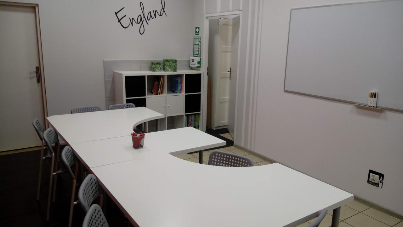 Jasna sala lekcyjna z białymi ścianami, długim, białym stołem w kształcie litery U otoczonym szarymi krzesłami, białą tablicą i regałem z książkami, z napisem 'England' na ścianie i naklejką...