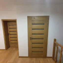 Dwie drewniane drzwi wewnętrzne z poziomymi, czarnymi wstawkami w nowym domu, widoczne z perspektywy klatki schodowej z drewnianą balustradą.