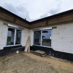 Fragment surowego budynku z dwoma oknami z częściowo opuszczonymi roletami zewnętrznymi, widoczne materiały budowlane i paleta na ziemi.