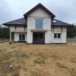 Surowy stan domu jednorodzinnego z zamontowanymi oknami PCV w kolorze antracytowym i drzwiami tarasowymi, widoczne bloczki betonowe i wstępne ocieplenie wokół okien, teren wokół domu w trakcie...