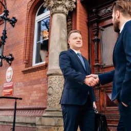 Dwóch mężczyzn w garniturach ściska sobie dłonie przed budynkiem sądu z kolumną i tablicą informacyjną