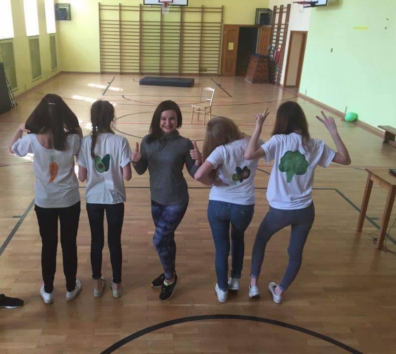 Grupa pięciu osób w sali gimnastycznej, cztery z nich ubrane w białe koszulki z nadrukami warzyw (marchewka, awokado, czosnek, brokuł), trenerka w szarej bluzie i legginsach, wszyscy uśmiechnięci...