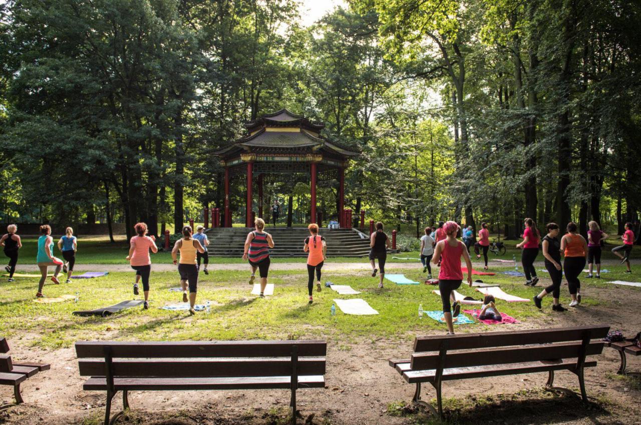 Grupa kobiet ćwiczy na trawie w parku przed orientalną pagodą, z widocznymi matami do ćwiczeń i ławkami w pierwszym planie.