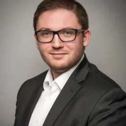 Portret mężczyzny w okularach i ciemnym garniturze na szarym tle, uśmiechnięty, z delikatnym zarostem.