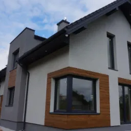 Nowoczesny dom jednorodzinny z szarą elewacją i fragmentami wykończonymi panelami imitującymi drewno, czarne okna i dachówka, widok z zewnątrz w pochmurny dzień.