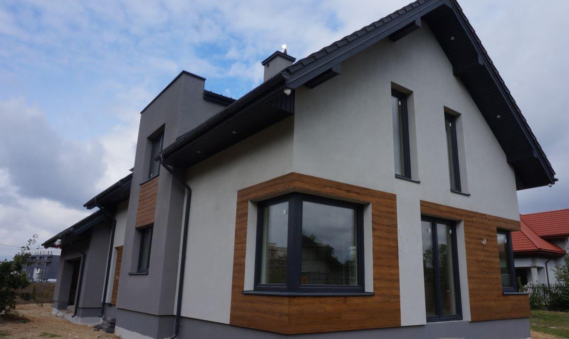 Nowoczesny dom jednorodzinny z szarą elewacją i fragmentami wykończonymi panelami imitującymi drewno, czarne okna i dachówka, widok z zewnątrz w pochmurny dzień.