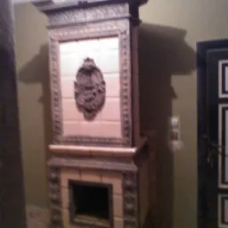 Stary, ozdobny piec kaflowy z dekoracyjnymi elementami i ornamentami, w tle ściana i fragment drzwi.