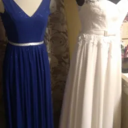 Dwa eleganckie, długie stroje wieczorowe: granatowa suknia z błyszczącymi aplikacjami i srebrnym paskiem oraz biała suknia z koronkową górą i paskiem, prezentowane na manekinach.