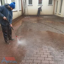Czyszczenie kostki brukowej myjką ciśnieniową przez dwóch pracowników w odzieży roboczej, widoczny brud i woda na powierzchni, drabina oparta o ścianę budynku w tle, logo firmy w rogu.