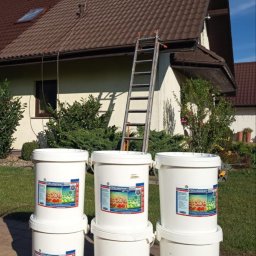 ANTON-FARBY ŁUKASZ ANTCZAK - Pracownik na dachu podczas aplikacji powłoki odbijającej ciepło. U dołu widoczne białe wiadra z etykietą 'Coolingpaint Roof' i drabina oparta o budynek.