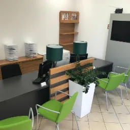 Wnętrze biura z recepcją, zielonymi krzesłami i rośliną w białej donicy, widoczne segregatory z dokumentami.