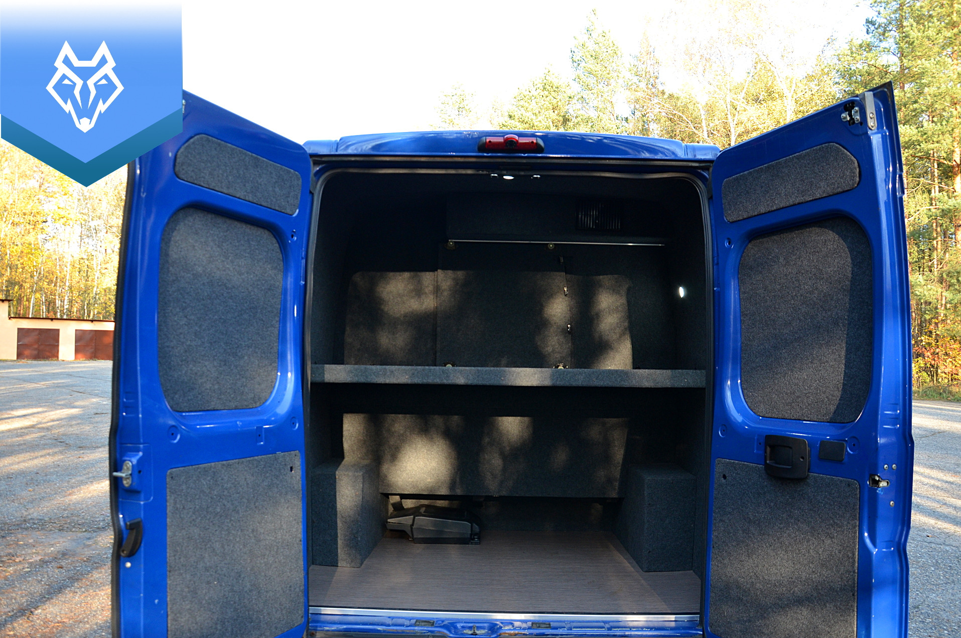 Wnętrze niebieskiego busa Peugeot Boxer VIP z otwartymi tylnymi drzwiami, wykończone szarą tapicerką i półkami, widok od tyłu na las w tle.