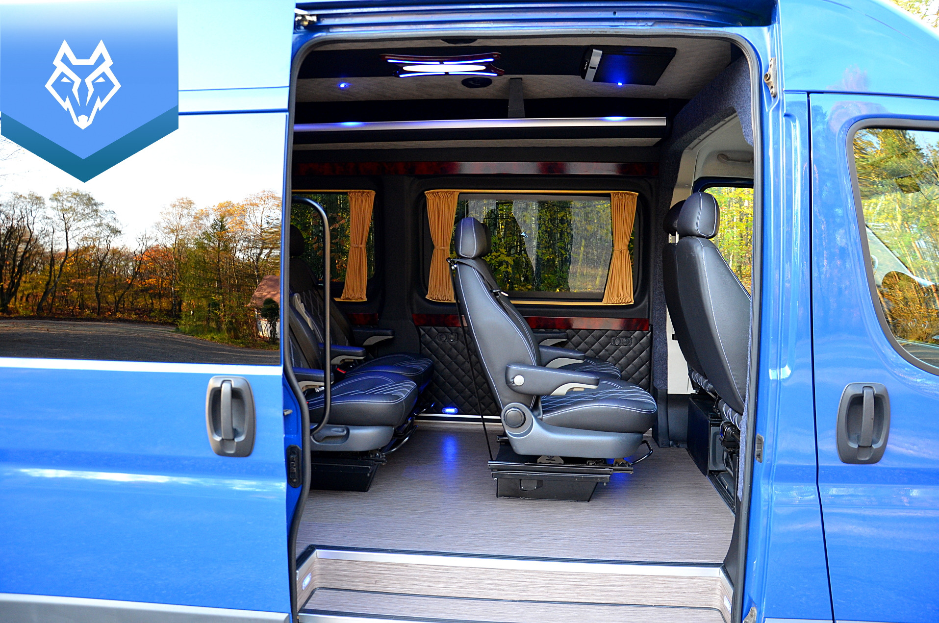 Wnętrze niebieskiego busa VIP z luksusowym wykończeniem, skórzanymi fotelami z białymi przeszyciami, drewnianą podłogą i zasłonkami w oknach.