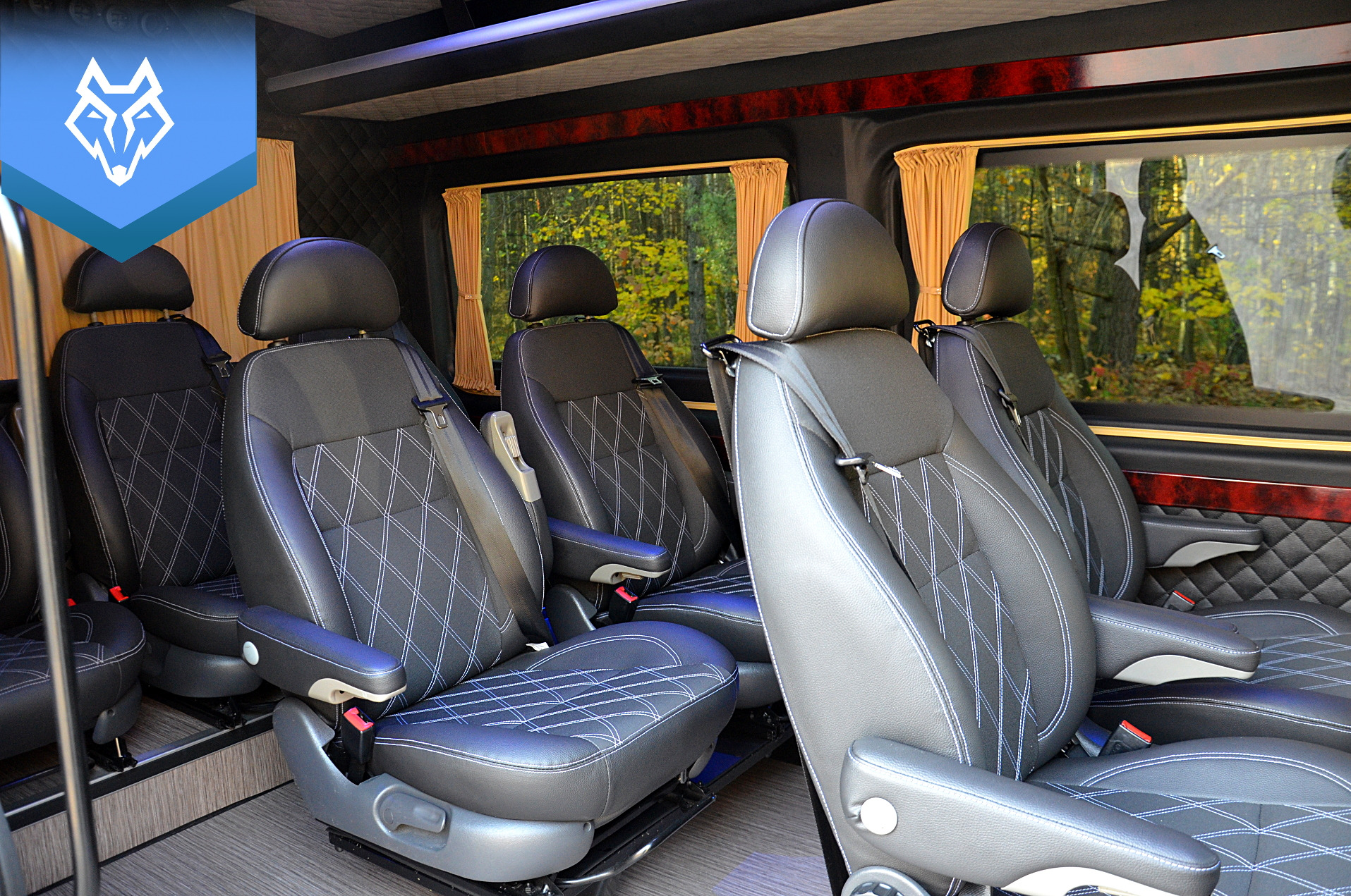 Wnętrze luksusowego busa VIP z czarnymi skórzanymi fotelami z białymi przeszyciami, widok na las za oknem z zasłonami.