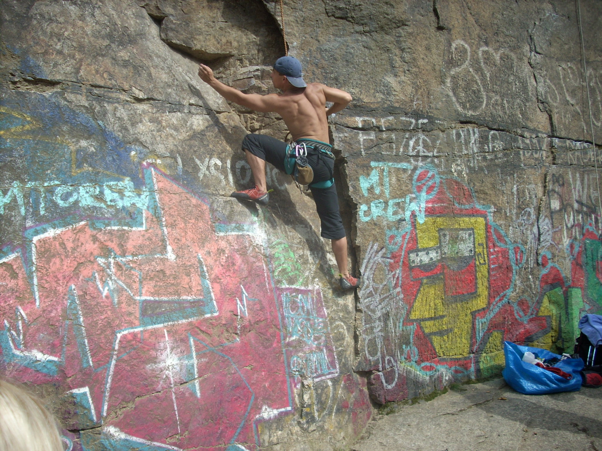 Mężczyzna bez koszulki, w czapce, wspina się na graffitti-zdobioną skałę, używając liny i uprzęży, z torbą sprzętu w pobliżu.