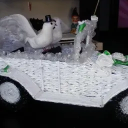 Ręcznie robiony kosz prezentowy w kształcie białego samochodu ślubnego z wikliny, ozdobiony białymi gołębiami w cylindrze, tiulem, sztucznymi różami i figurką krasnala.