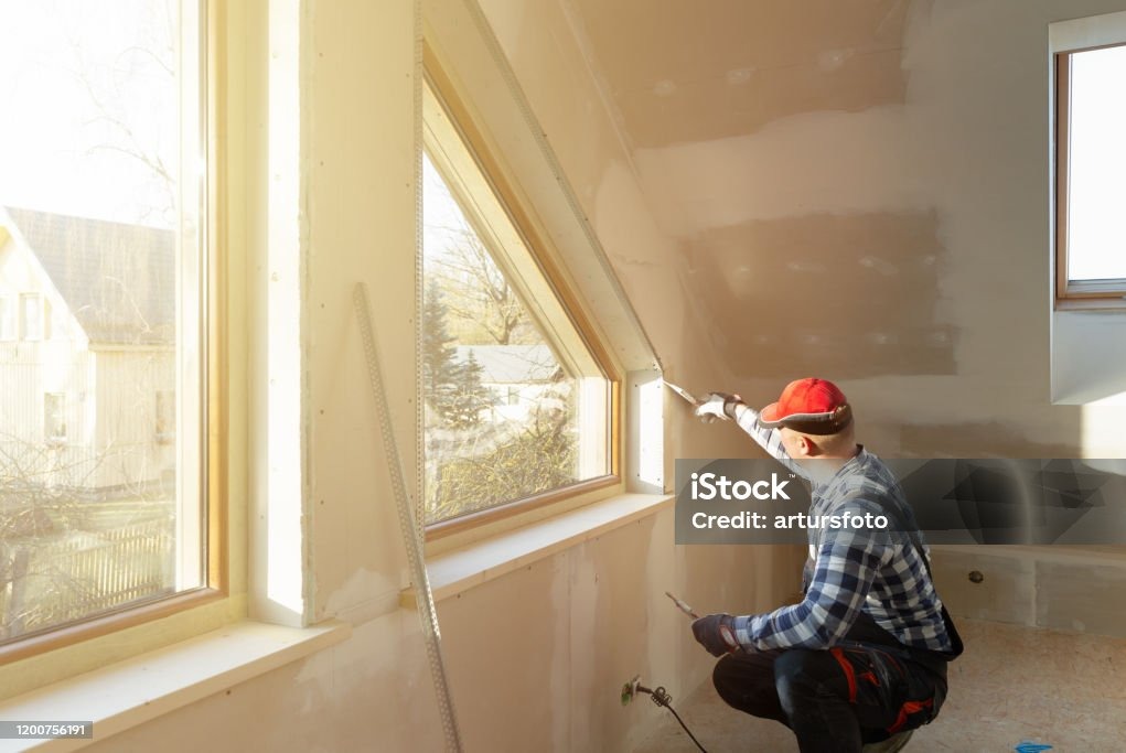 Praca przy gładzi: mężczyzna w czerwonej czapce szpachluje ścianę przy oknie na poddaszu. Widoczne narzędzia, profil aluminiowy, częściowo wyszpachlowana ściana.