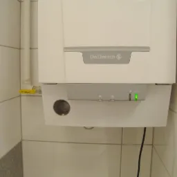 Biały, wiszący kocioł gazowy marki De Dietrich na tle białych płytek. Widoczny żółty zawór gazowy i zielona dioda sygnalizacyjna.