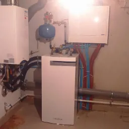 Zestaw urządzeń grzewczych w kotłowni: dwa białe kotły, zbiornik wyrównawczy, instalacja z rur miedzianych i plastikowych w kolorze czerwonym i niebieskim.