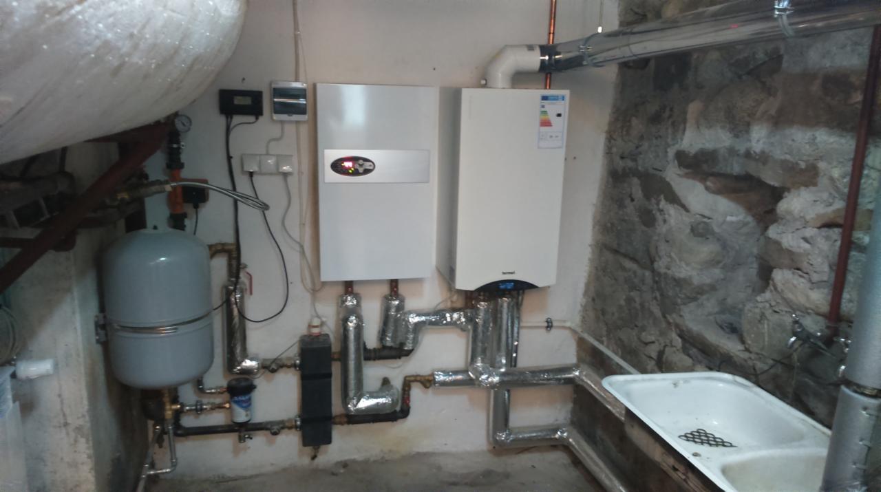 Dwa białe kotły grzewcze zamontowane na ścianie w piwnicy z kamiennymi elementami, widoczne rury w izolacji, zbiornik wyrównawczy i stara umywalka.