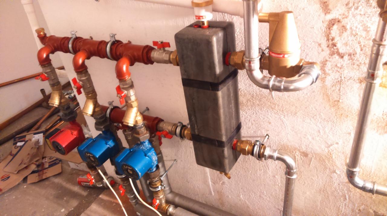 Rozbudowana instalacja hydrauliczna z czerwonymi rurami, niebieskimi pompami obiegowymi i szarym zbiornikiem wyrównawczym na tle surowej ściany.