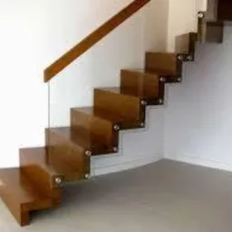 Nowoczesne schody drewniane z przezroczystymi szklanymi balustradami, mocowane na stalowych wspornikach, oświetlone naturalnym światłem padającym z boku, widoczne na tle białej ściany i szarej...