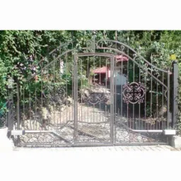 Kuta, dwuskrzydłowa brama wjazdowa z furtką pośrodku, ozdobiona secesyjnymi ornamentami, z automatyką do otwierania, na tle zieleni.