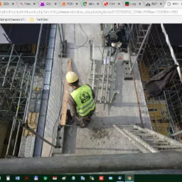 Pracownik budowlany w żółtym kasku i kamizelce z logo firmy, stojący na szalunku zbrojeniowym, widok z góry na placu budowy z rusztowaniami i drabiną.