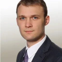 Portret biznesowy młodego mężczyzny w ciemnym garniturze w prążki i fioletowym krawacie na jasnym tle.