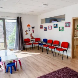 Jasna sala lekcyjna z rzędem czerwonych krzeseł z podkładkami, białym stołem otoczonym kolorowymi taboretami, dekoracjami ściennymi z postaciami z kreskówek i rośliną doniczkową przy oknie.