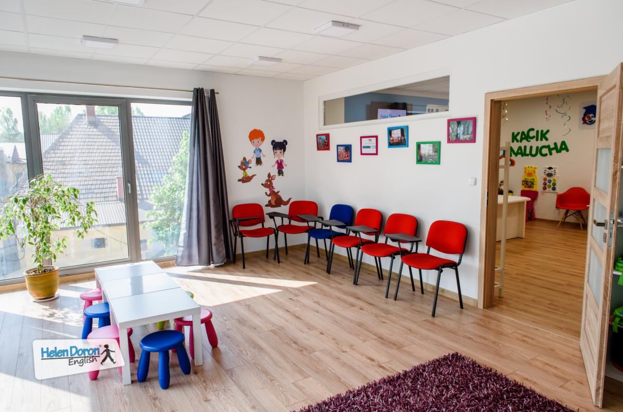 Jasna sala lekcyjna z rzędem czerwonych krzeseł z podkładkami, białym stołem otoczonym kolorowymi taboretami, dekoracjami ściennymi z postaciami z kreskówek i rośliną doniczkową przy oknie.