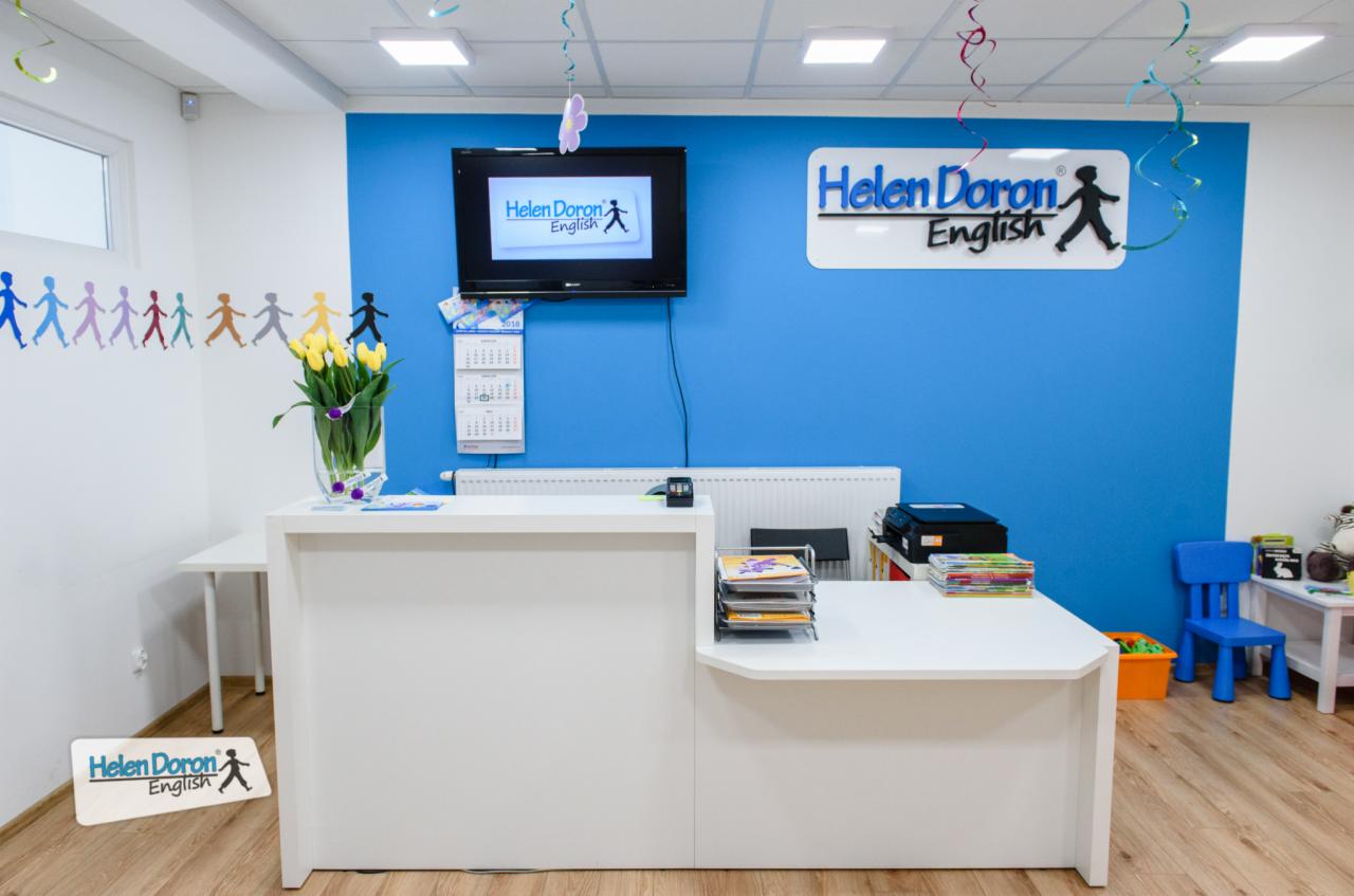 Recepcja szkoły językowej Helen Doron English z dekoracjami, kwiatami w wazonie i logo na ścianie oraz na podłodze.