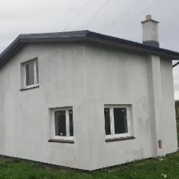 Niedokończony, otynkowany budynek z dwoma oknami na każdej ścianie, kominem i widoczną linią wysokiego napięcia w tle.