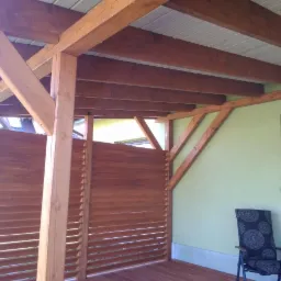 Zadaszona konstrukcja z drewna z ażurowymi ścianami, widoczne elementy konstrukcyjne z belek, krzesło na tarasie, okno z doniczką na ścianie budynku.