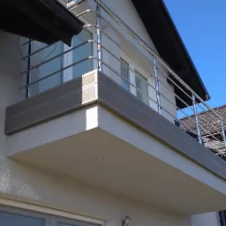 Balkon z chromowanymi barierkami na tle błękitnego nieba i fragmentu dachu sąsiedniego budynku.