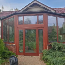Zewnętrzny widok drewnianej oranżerii z przeszklonymi ścianami i drzwiami, przylegającej do domu z czerwoną dachówką, otoczonej zielenią i brukowanym chodnikiem.