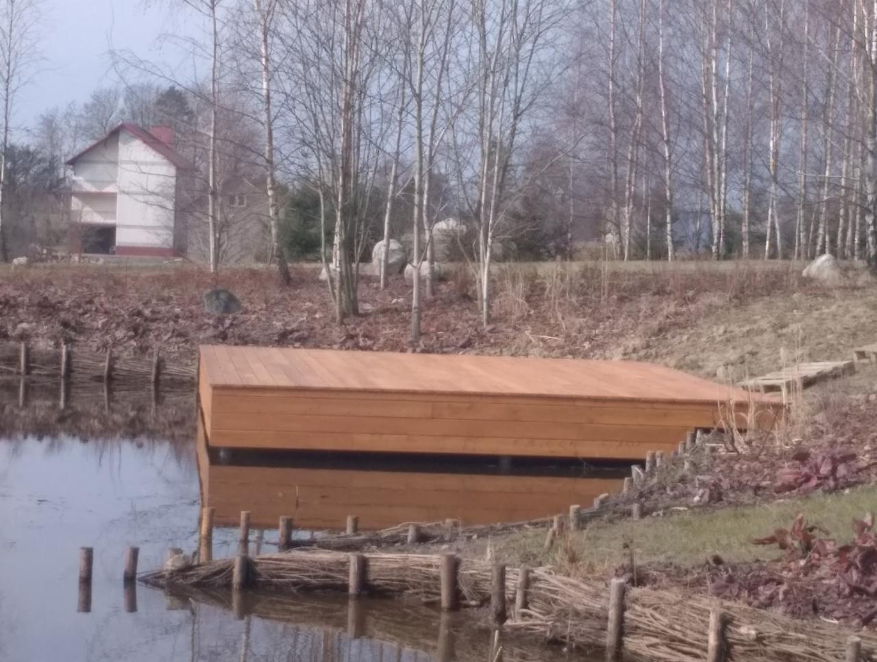 Drewniana platforma nad wodą, z widocznym odbiciem w tafli jeziora, otoczona naturalnym brzegiem i brzozowym lasem w tle. Niska zabudowa z czerwonym dachem widoczna w oddali.