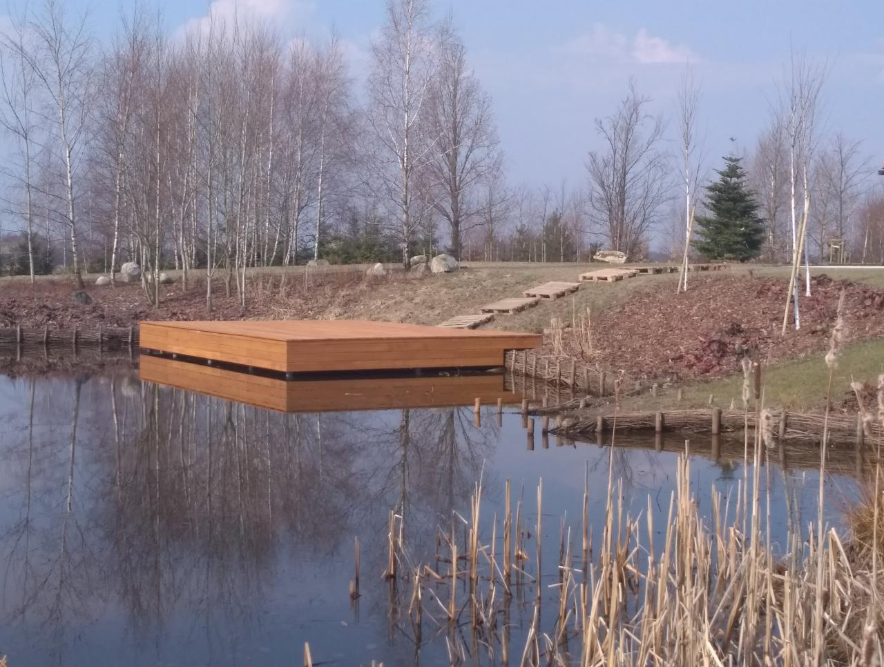 Drewniany pomost na stawie, odbijający się w wodzie, z widoczną ścieżką z drewnianych palet prowadzącą w górę skarpy.