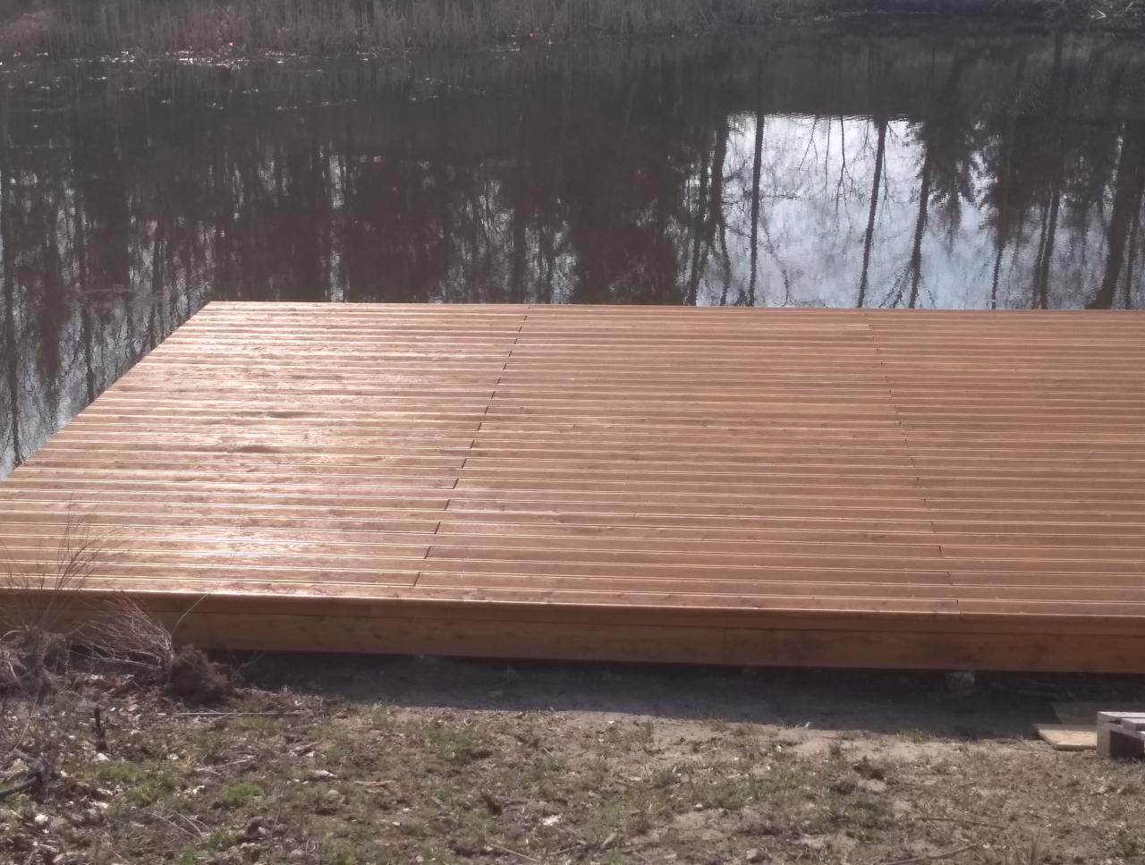 Nowo zbudowany drewniany podest nad wodą, z widocznymi śladami ziemi i trawy na brzegu, w tle odbicie drzew w spokojnej tafli wody.